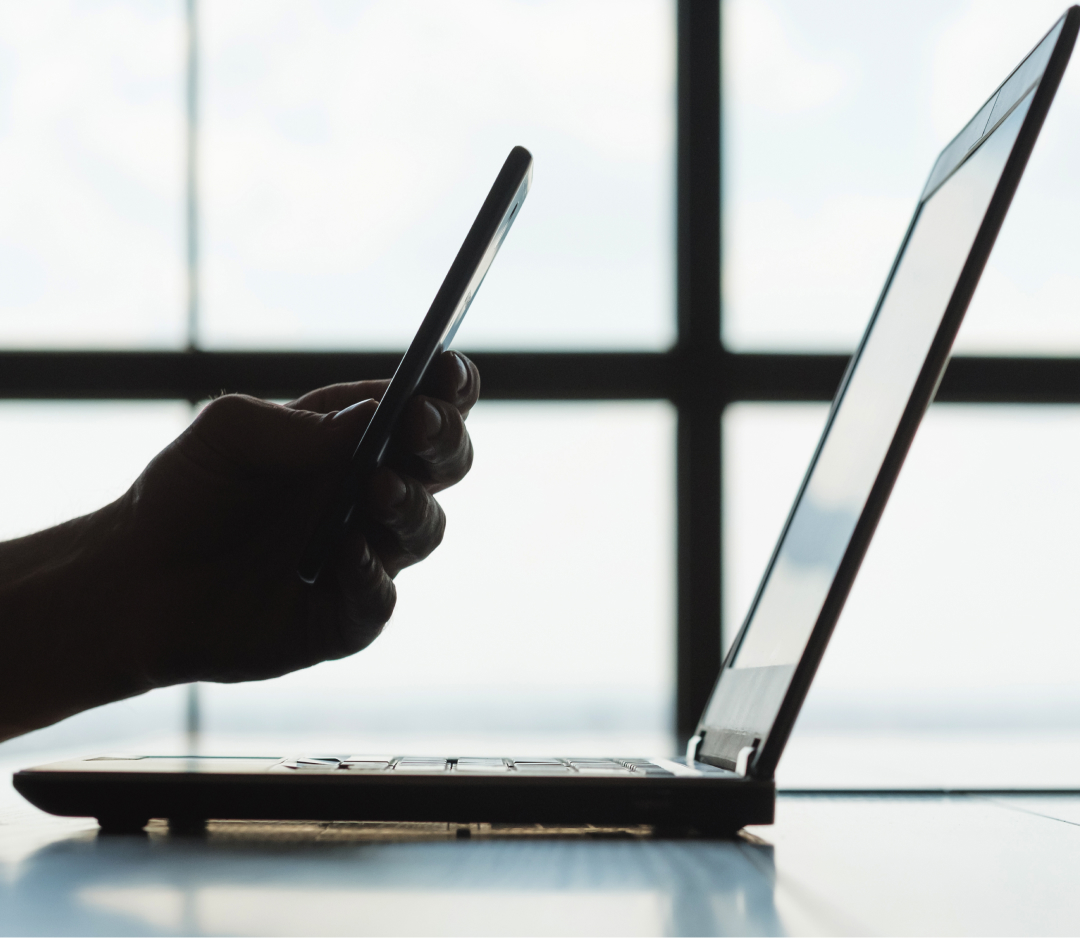 A silhouette of a hand holding a smartphone integrates seamlessly with the identity of an open laptop on a desk.