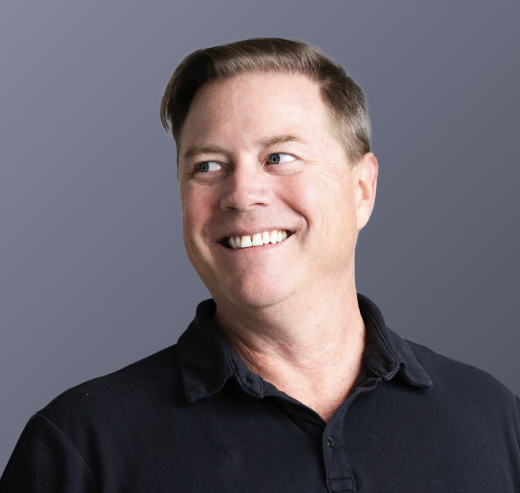 A smiling man with short, light brown hair wearing a dark polo shirt looks slightly to his right against a plain grey background, embodying the professionalism found in Forrester TEI: Maverics Identity Orchestration.